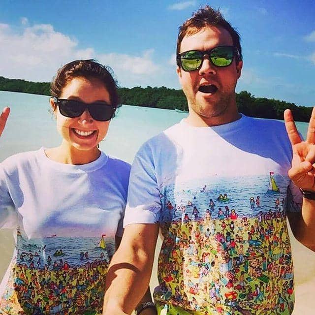 Smiling couple in matching illustrated beach t-shirts, sunglasses, posing by a lake