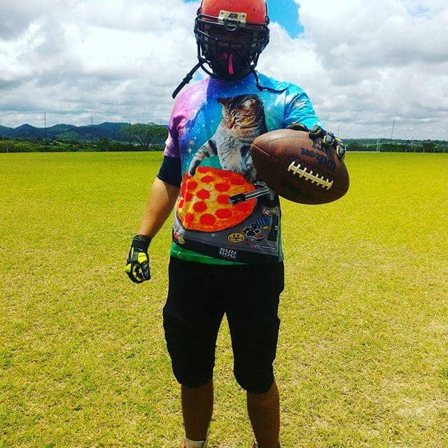 Football player on field wearing cat pizza galaxy shirt, holding football outdoors