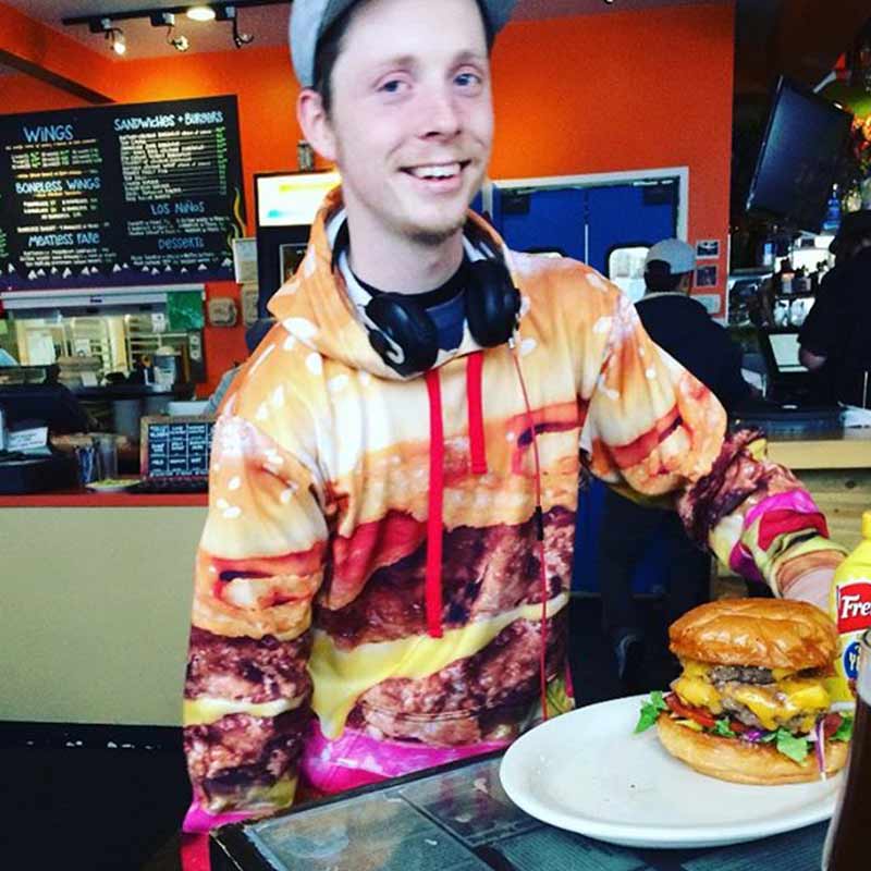 Person wearing a colorful hoodie with a burger design, sitting at a table with a large burger in front of them.