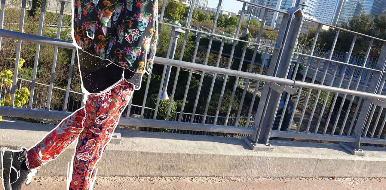 Person wearing colorful pants and a patterned top on a bridge with metal railings and greenery.