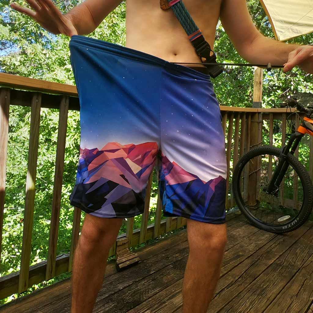 Poly Mountain Men's Gym Shorts - The sharp, triangular tessellation of the "dusky magenta peaks" (#B65C72) contrasting against the smooth "cosmic cobalt" (#2C4A7C) gradient, evoking the background rendering of 32-bit adventure games.